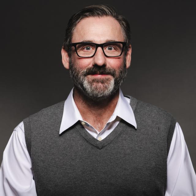 Mark Noble wearing a shirt, sweater vest, and glasses smiling for his headshot with a black background.